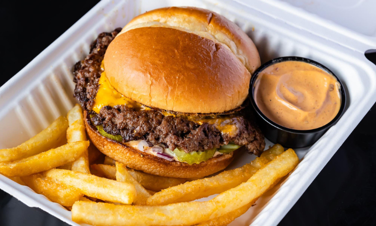 The OG Smash: double smashed beef patties with melted American cheese, pickles, and onions on a pretzel bun, served in a takeout box with crispy fries and SMASH sauce.